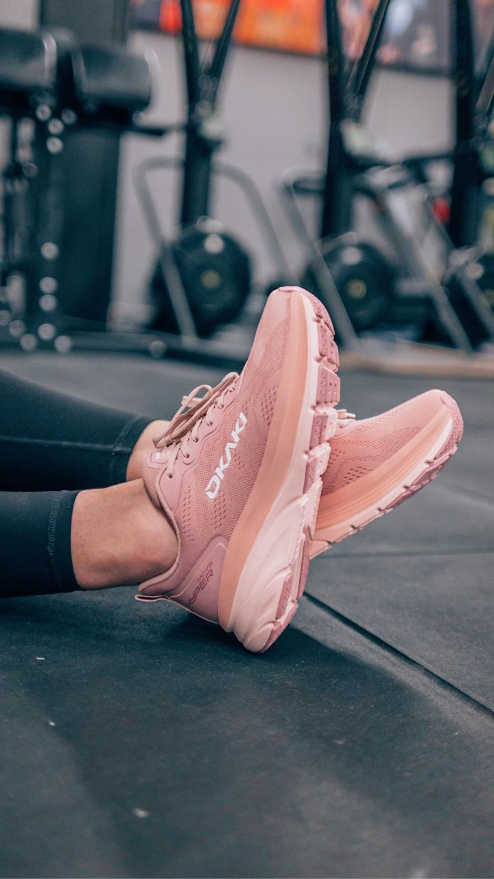 Rosa Ukaki träningsskor med texten "MOVE HYPER" på sidan, bärs av en person som sitter på golvet i ett gym.
