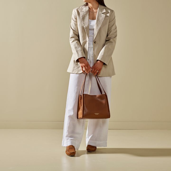 Kvinne som holder en brun skinnveske, iført beige blazer, hvit topp og vide hvite bukser, stående mot en beige vegg.