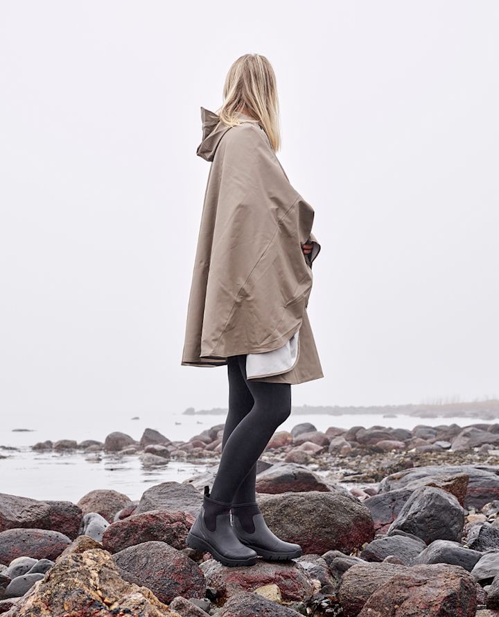 Kvinna med blont hår står på stenar vid havet iförd beige regnponcho, svarta strumpbyxor och svarta stövlar en mulen dag.