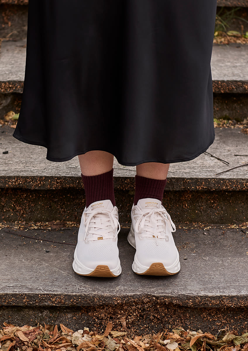 Vita Okaki Mile sneakers med gummisulor och vinröda strumpor under en svart kjol, stående på stentrappa med höstlöv.