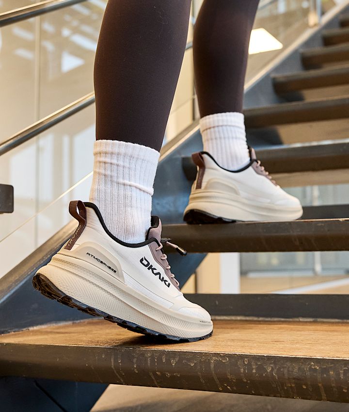 Person i svarta leggings och vita strumpor bär vita Okaki fritidsskor med svarta detaljer på en trätrappa.