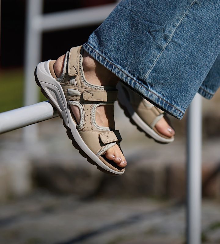 Beige sportsandaler med borrelåsremmer og hvit såle, brukt med blå jeans. Føttene hviler på et gelender utendørs.