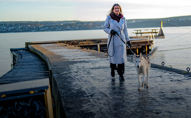 Kvinna i ljus kappa promenerar med en vit och grå hund på en brygga vid vattnet med stad i bakgrunden.