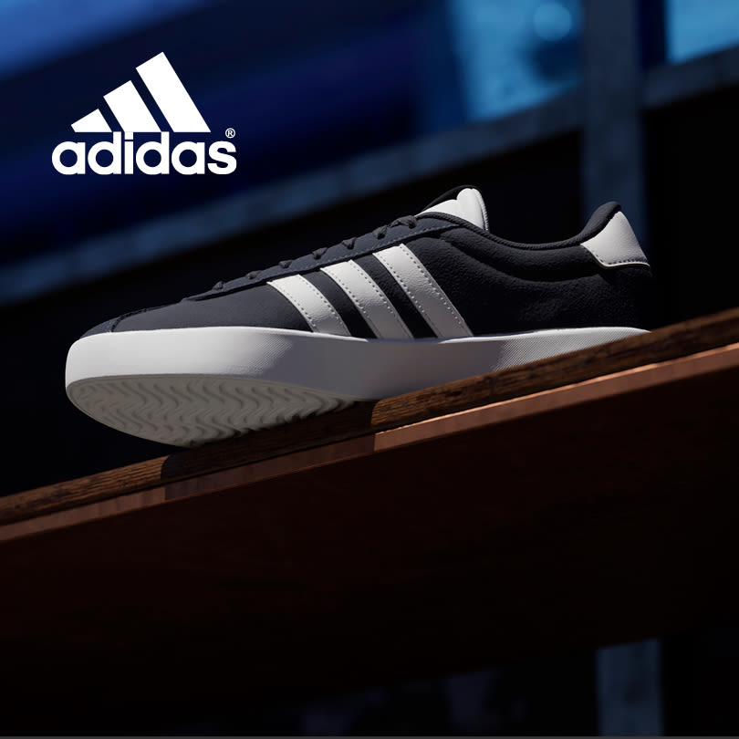 Svart Adidas sneaker med sølvgrå striper og hvit såle, plassert på en treoverflate mot blå bakgrunn med Adidas-logo.