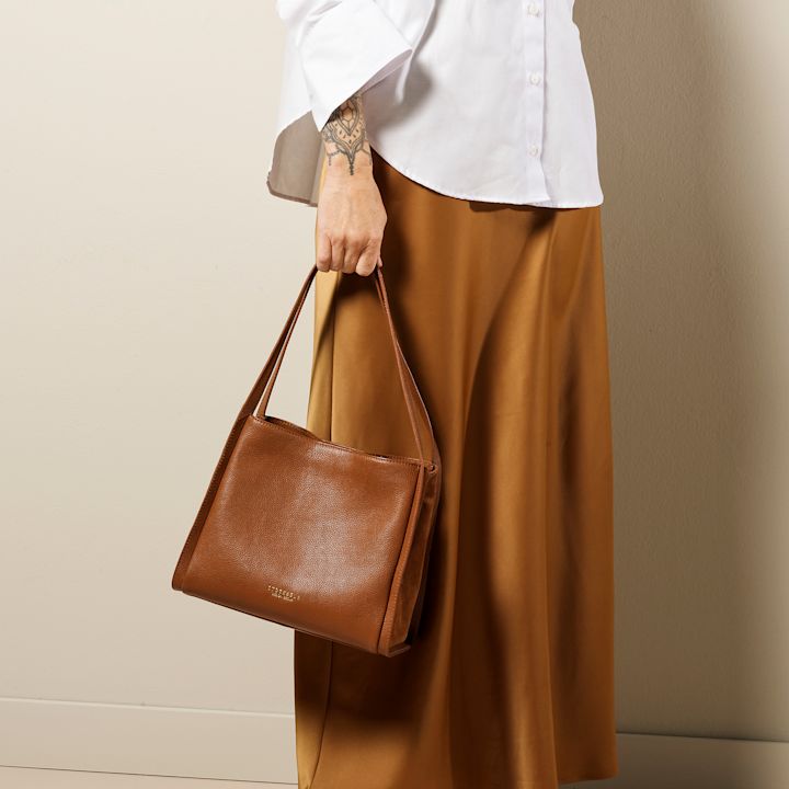 Person holder en brun skinnveske med to stropper, iført hvit skjorte og brun lang skjørt, stående foran en beige vegg.