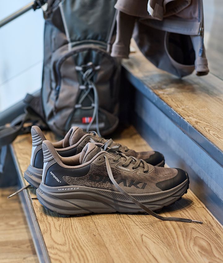 Fritidssko fra Okaki med Maxgrip-såle på tretrapp ved siden av en ryggsekk, designet for turer med robust såle og snøring.