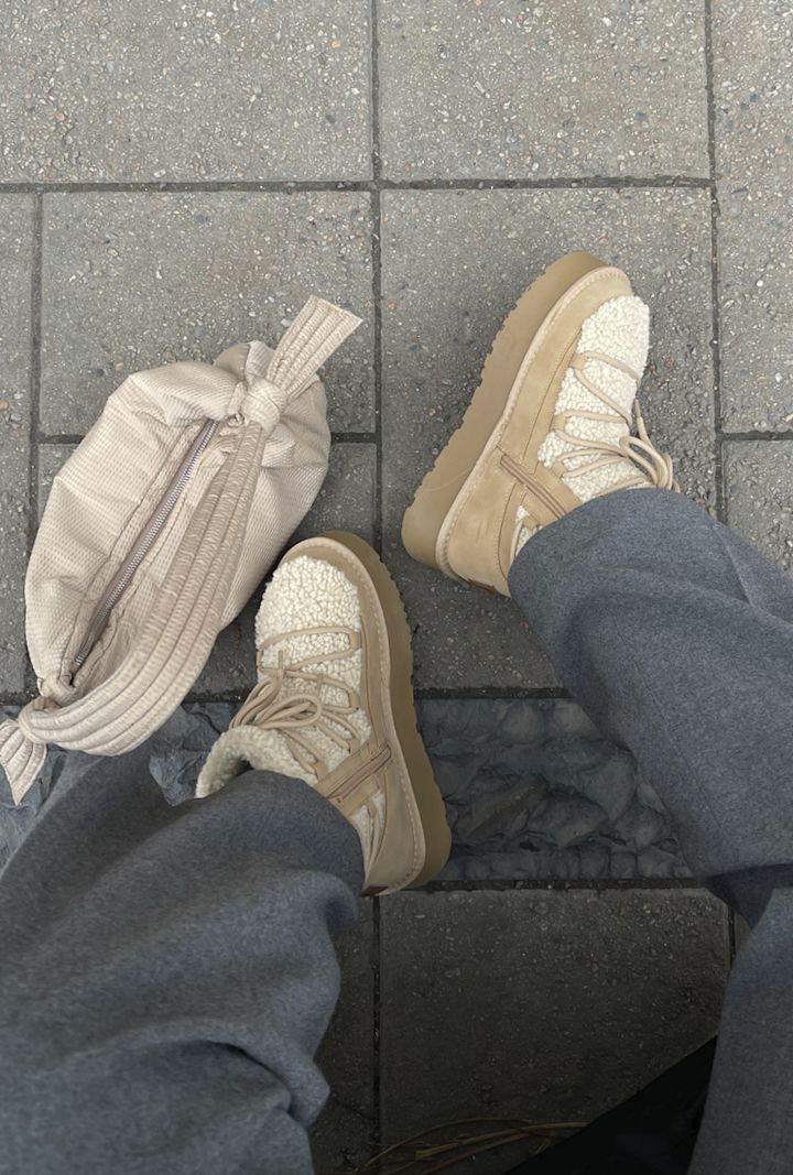 Beige vinterstövlar med snörning och dragkedja på fötterna på en person som bär grå byxor, bredvid en beige väska på en kullerstensbelagd gata.
