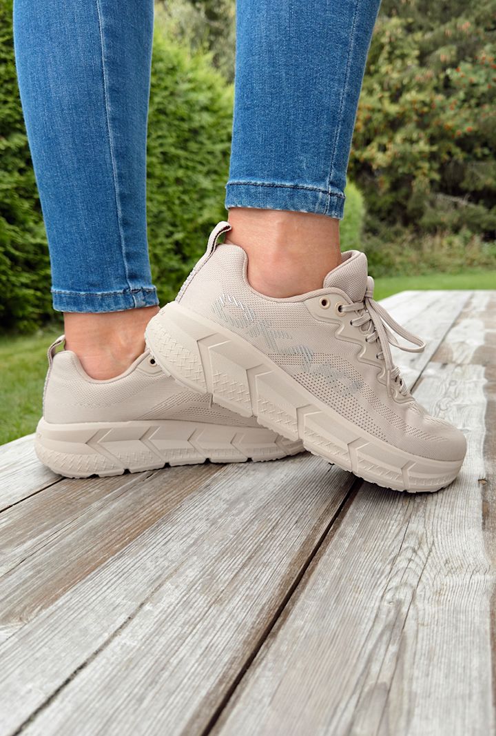 Beige joggesko med tykke såler som bæres av en person i blå jeans, stående på et tredekke utendørs med grønn busk i bakgrunnen.