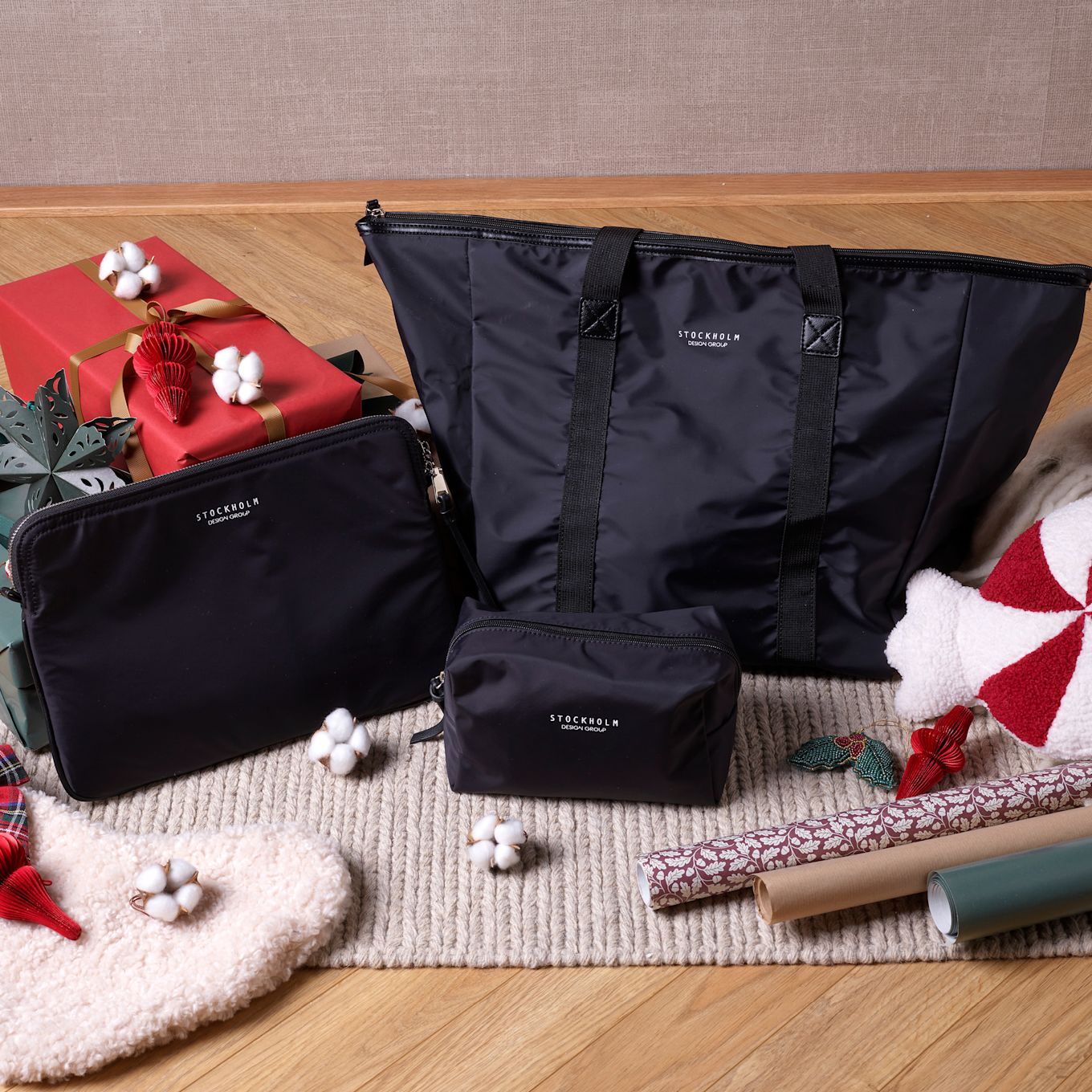 Svarta Stockholm-väskor i olika storlekar med julklappar, bomullsblommor och presentpapper på en stickad matta.