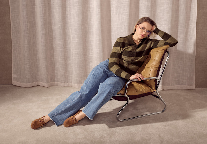 Person i stripete genser og jeans sitter i en stol med beige pute, iført brune semsket mokasiner med snøring.