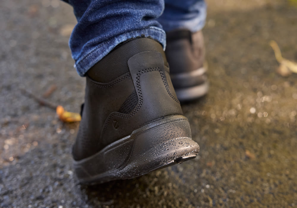Mørkebrune vanntette støvler med vanndråper på sålen, brukt med mørkeblå jeans på våt bakke.
