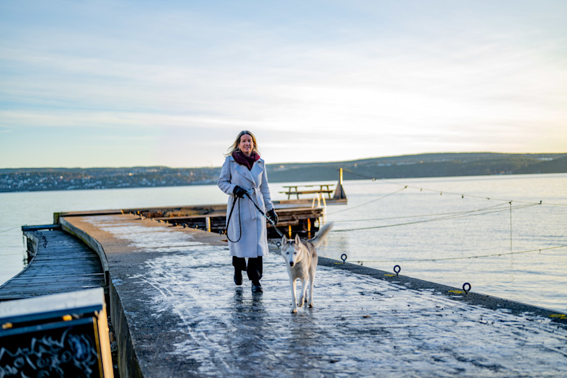 Kvinne i lys kåpe går med hund på en snødekt brygge ved vannet med fjell i bakgrunnen.