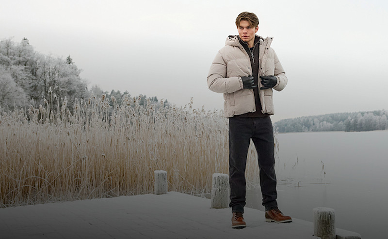 Ung man som tar på seg en ljus vinterjacka medan han står på en snötäckt brygga vid en sjö med frosttäcka träd och vass i bakgrunden.