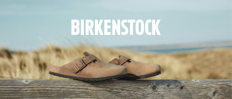 Logo: BIRKENSTOCK over et par brune Birkenstock-sandaler som hviler på et trestykke med tørt gress og blå himmel i bakgrunnen.