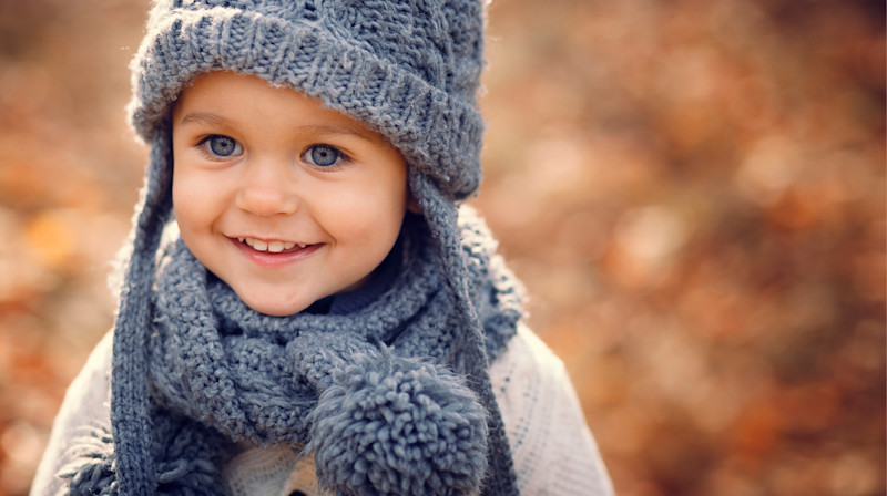 Litet barn med blå ögon ler och har på sig grått stickat mössa och halsduk i höstlandskap med suddig bakgrund.