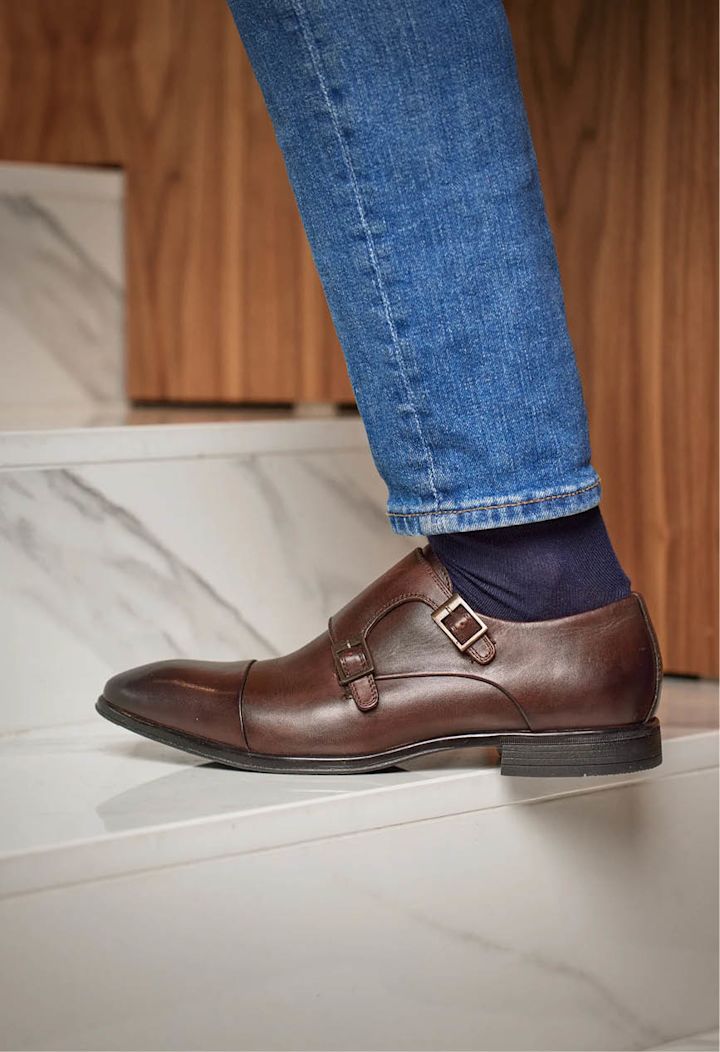 Brun lærsko med spenne på foten til en person som går opp hvite marmortrapper med blå jeans og mørk sokk.