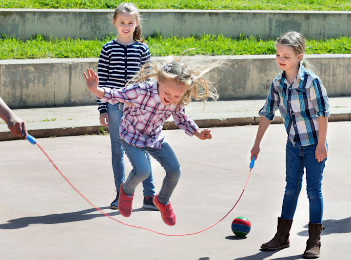 Tre barn leker hopprep ute på en betongyta med en grässluttning i bakgrunden, ett barn hoppar över repet medan två andra svingar det.