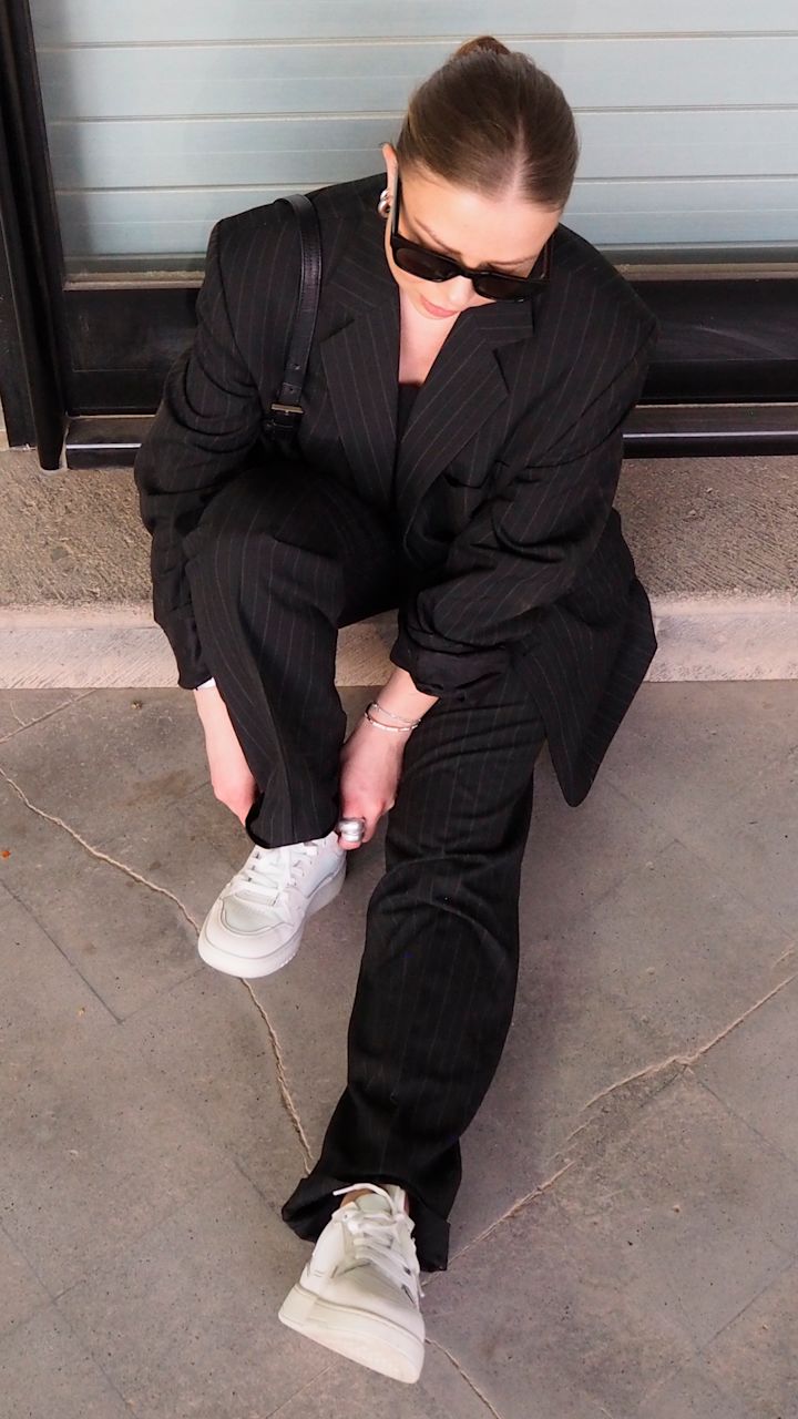 Kvinna i svart stripete dress och solglasögon som sitter på marken och knyter vita joggingskor utanför en byggnad.