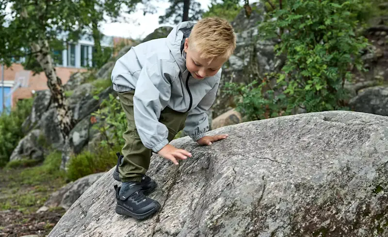 Barn i grå jacka klättrar på klippa i naturen, bär svarta robusta vandringskängor med bra grepp.