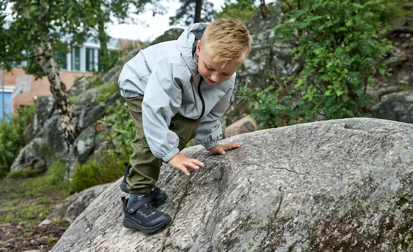 Barn i grå jakke klatrer på stein i naturen, iført svarte robuste tursko fra Okaki med Sympatex-membran.