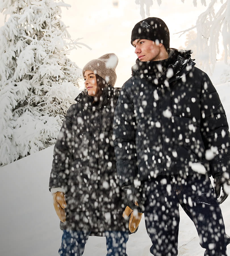 Par i svarta vinterjackor och stickade mössor promenerar tillsammans i snöfall bland snötäckta träd i vinterlandskap.