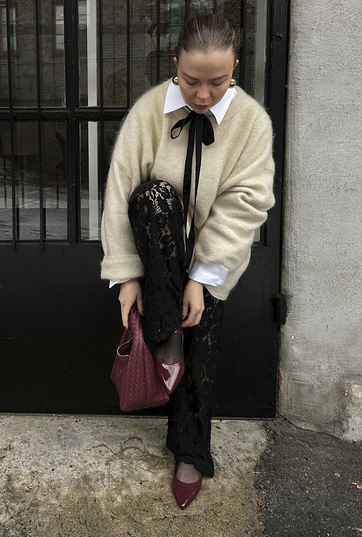 Kvinna bär en beige tröja med vit skjorta och svart rosett, böjer sig ner för att justera vinröda skor, håller en vinröd väska, framför en svart dörr och en grå vägg.