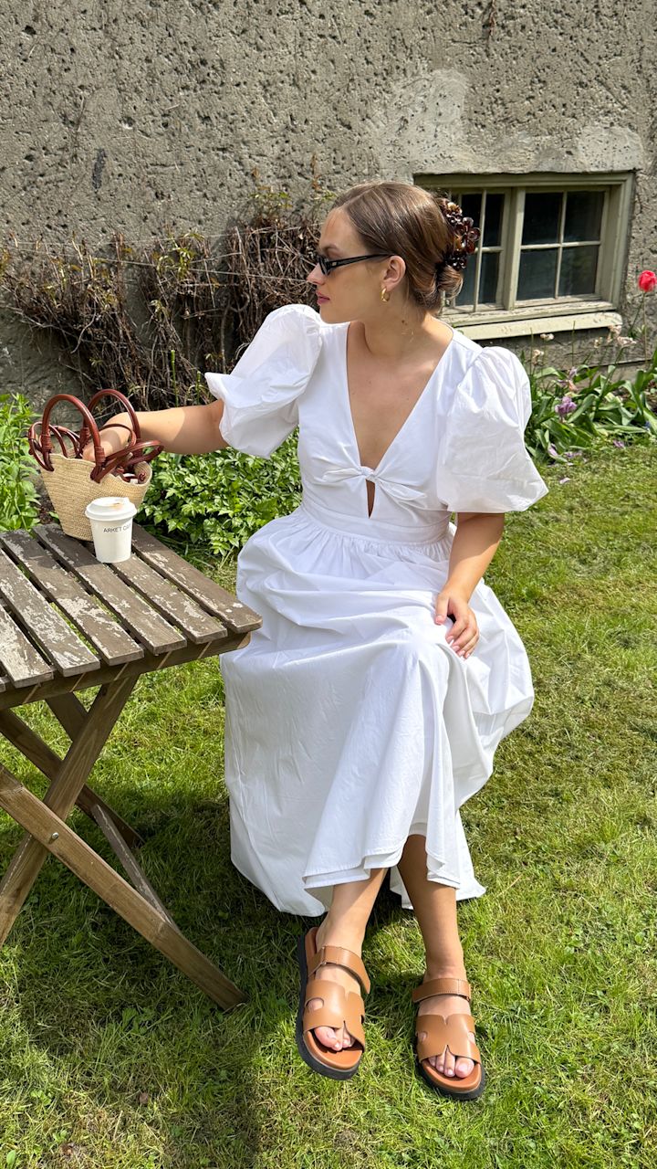 Kvinna i en vit klänning med puffärmar och bruna sandaler sitter på gräset vid ett träbord med en kaffekopp och en väska i en trädgård med blommor och en stenmur med ett fönster i bakgrunden.