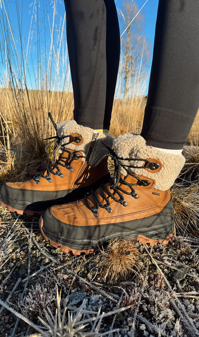 Bruna och svarta vinterstövlar med fleecefoder och snörning, bärs av en person i svarta tights, står på frosttäckt gräs i ett öppet, soligt landskap.