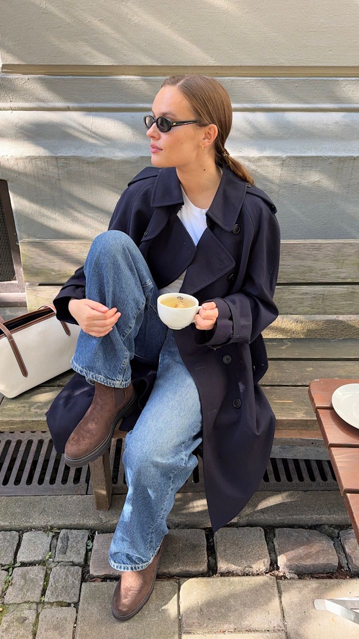 Lisa Alexandra i mörkblå kappa och jeans sitter på bänk med kaffekopp, bär bruna chelsea boots med robust sula i avslappnad stil.