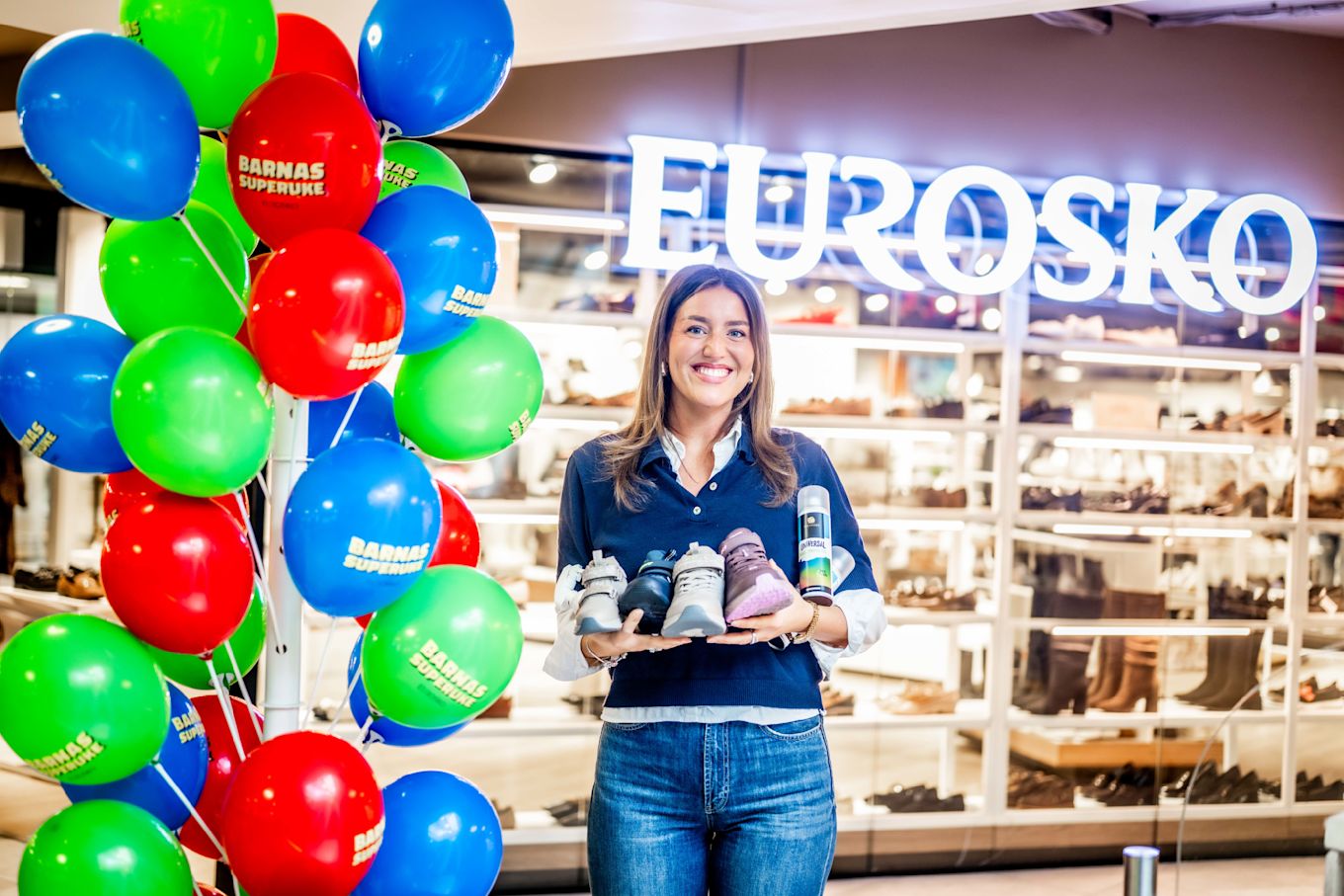 Smilende butikkansatt holder barnesko i ulike farger foran Eurosko-butikken, omgitt av fargerike ballonger.