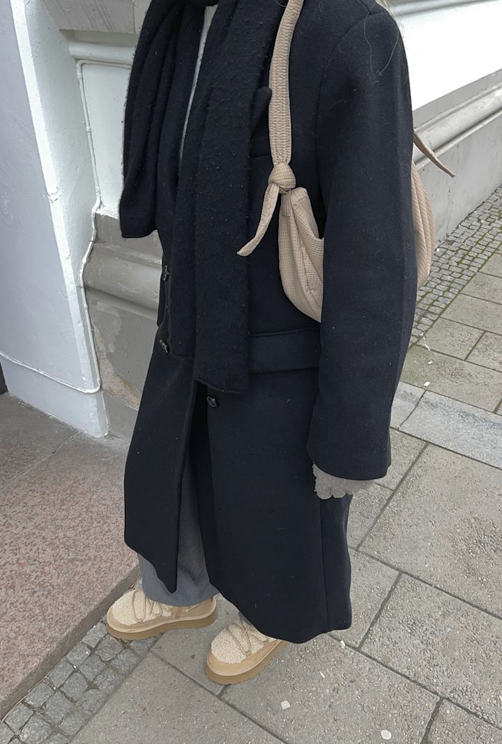 Person klädd i svart kappa och halsduk, grå byxor och beige skor, med en beige väska över axeln, står på trottoaren vid en byggnad.
