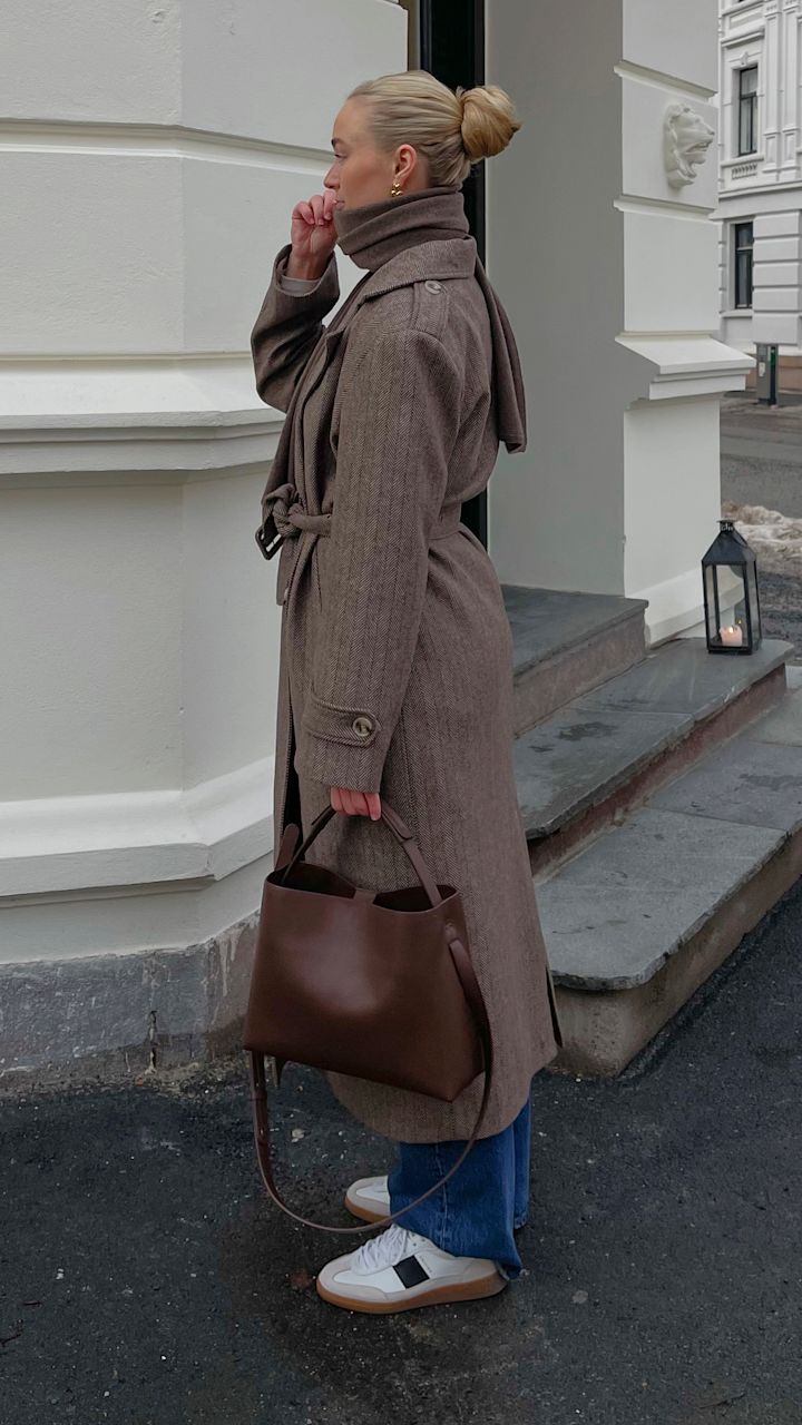 Kvinna med blont hår i knut, iklädd lång brun kappa, blå jeans och vita sneakers, håller en brun skinnväska och står vid en trappa utanför en vit byggnad.