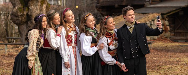 Gruppe i tradisjonelle norske bunader med fargerike hodeplagg tar selfie utendørs i vårlandskap. Bunadstilbehør fra Noma Norge. Foto: Noma Norge