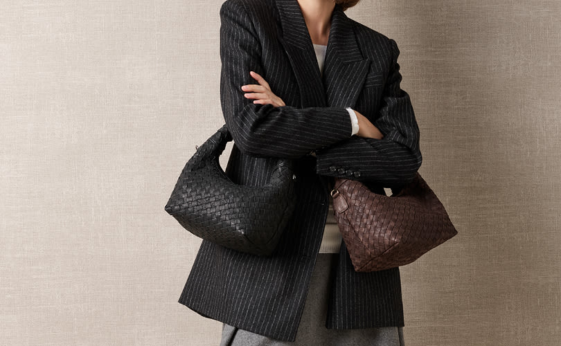 Person i svart randig kavaj med armarna i kors, bärandes en svart och en brun flätad väska mot beige bakgrund.