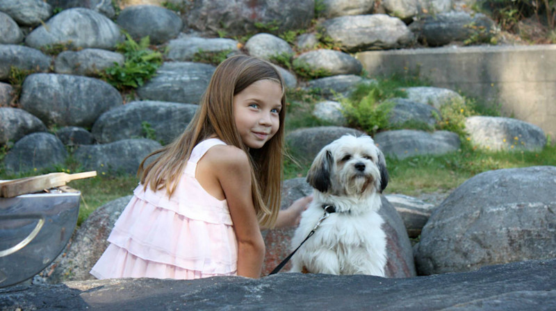 Flicka i rosa klänning som klappar en liten vit och svart hund utomhus med stenar och gräs i bakgrunden.