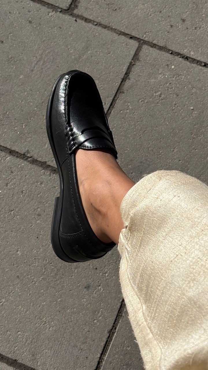 Svart klassisk loafer i läder med dekorativa sömmar på en stenbelagd yta, buren med ljusa byxor.