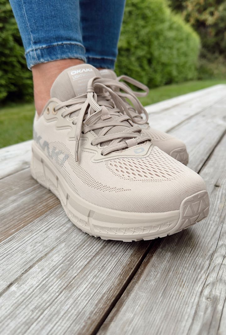 Beige Okaki löparskor med snörning på en person som står på en träplattform utomhus med grönt gräs och buskar i bakgrunden.