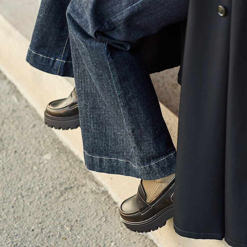 Svarta loafers med kraftig platåsula under vida jeans, beige strumpor syns mellan skorna och jeansen.