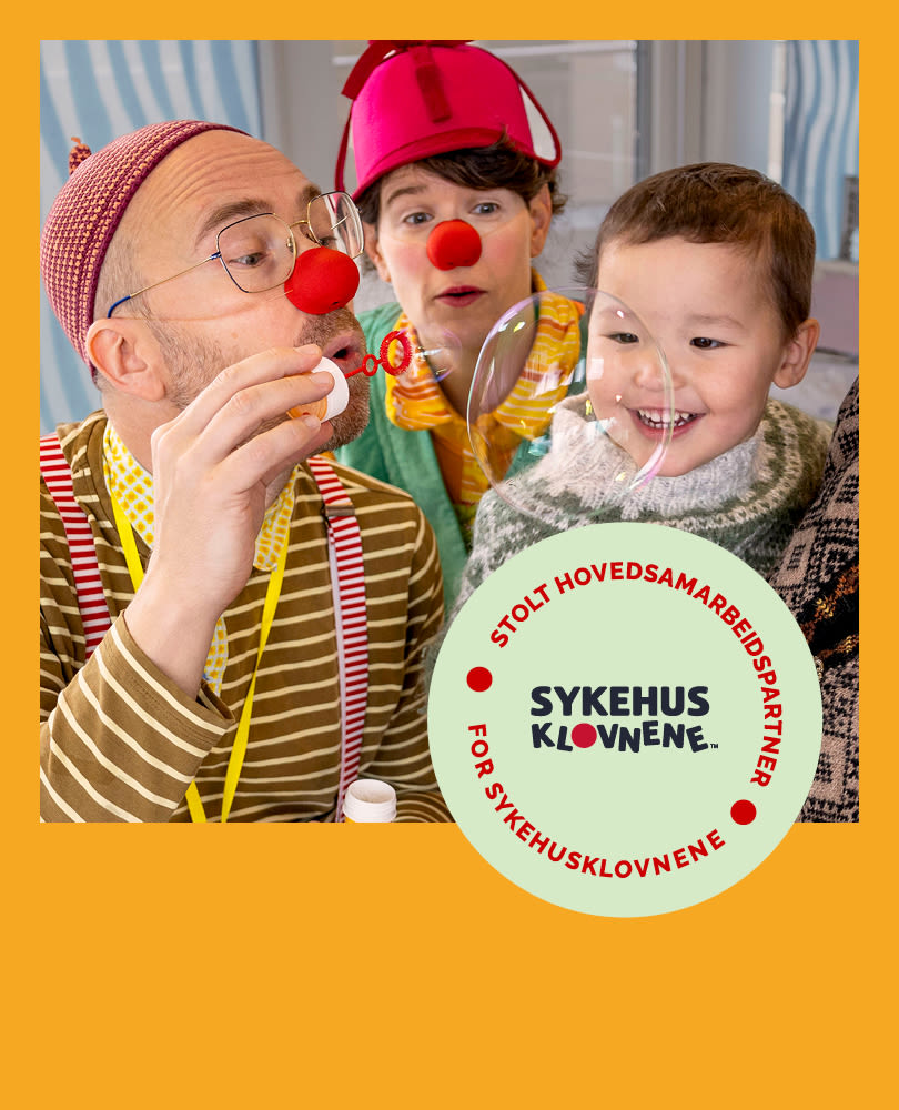 To klovner med røde neser underholder et smilende barn. Sykehusklovnene-logo vises i hjørnet på gul bakgrunn.