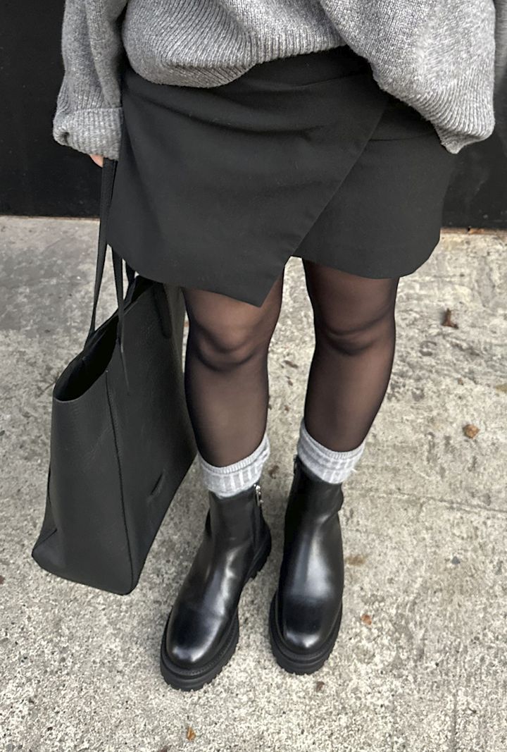 Person iförd svart kjol, svarta stövlar med gråa strumpor, och håller en svart väska, står på en grå trottoar.