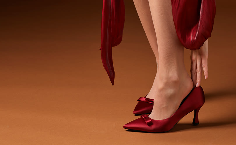 Eleganta röda pumps med spetsig tå och rosettdetalj, matchande en röd klänning mot brun bakgrund.