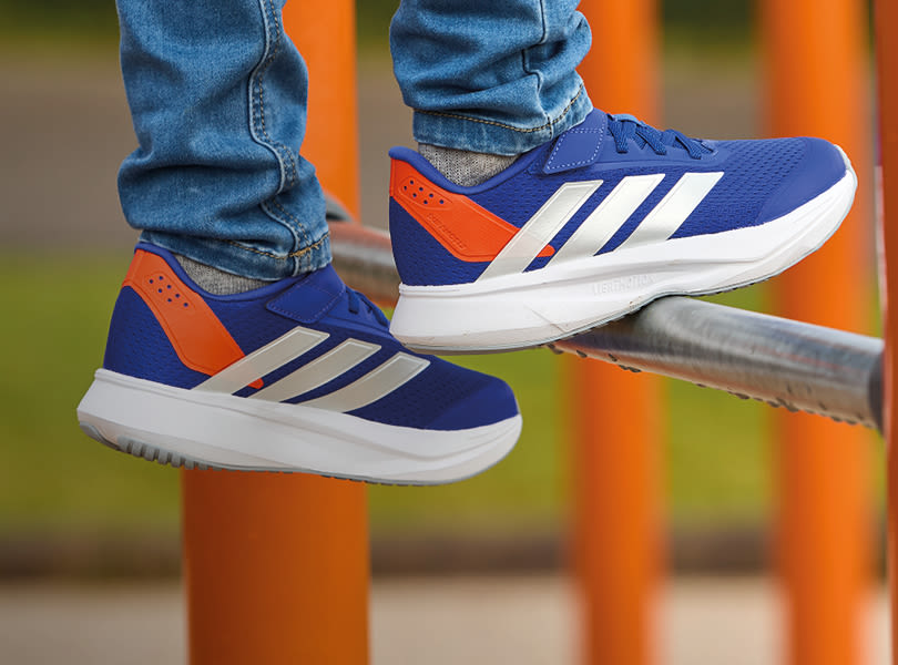 Blå Adidas løpesko med hvite striper og oransje detaljer, brukt av person i jeans som balanserer på metallrekkverk.