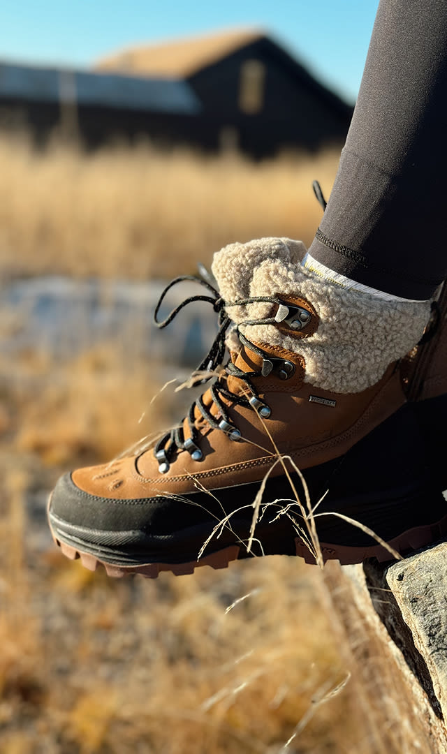 Brun og svart vinterstøvel med ullfôr som bæres på en fot i utendørs, gresskledd område med en bygning i bakgrunnen.