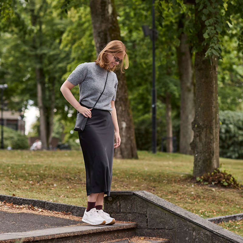 Kvinne iført grå genser, svart skjørt og hvite sko går ned en trapp utendørs i en park med trær og gress.