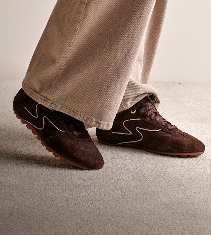 Mörkbruna mocka sneakers med beige vågmönster och gummisulor, parade med beiga byxor.