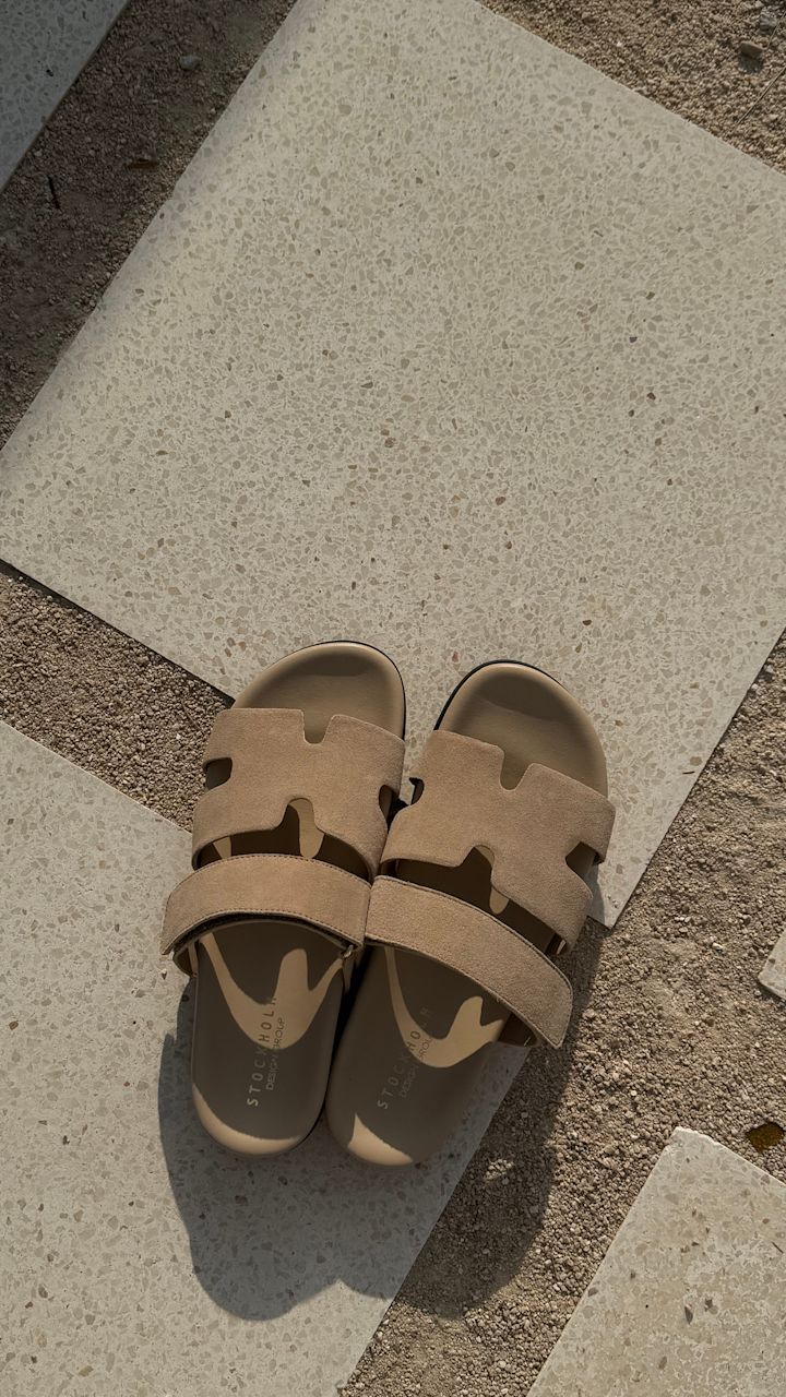 Beige semsket sandaler med H-formet design på oversiden fra Stockholm Design Group, plassert på en terrazzo-flisoverflate i nøytrale fargetoner.