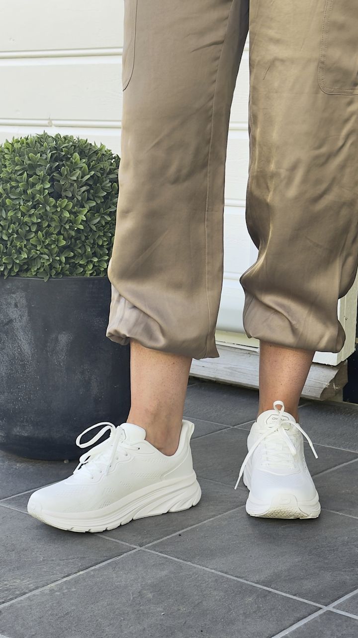 Hvite joggesko med snøring på en person iført beige bukser som står på grå flislagt gulv ved siden av en svart potte med grønn plante.