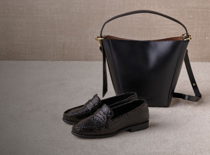 Svarta flätade loafers i läder bredvid en elegant svart läderväska från Stockholm Design Group med bruna detaljer mot en neutral bakgrund.
