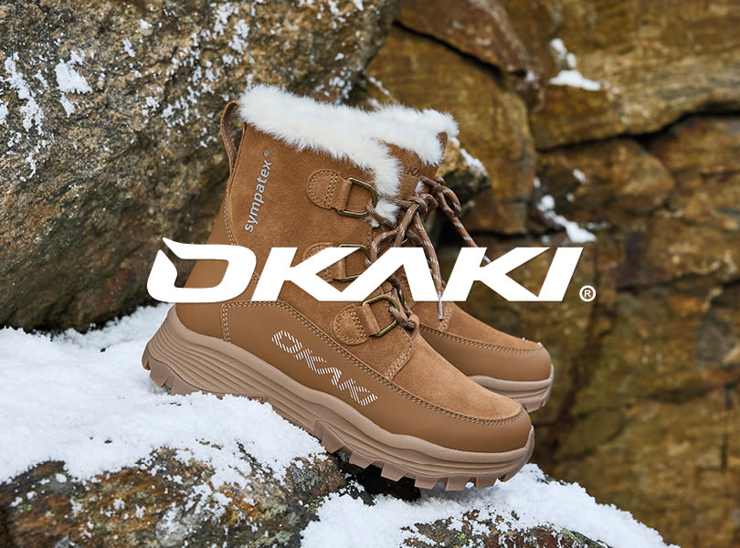 Bruna vinterkängor från OKAKI med vit pälskant och snörning, placerade på snötäckt klippa i vintermiljö.