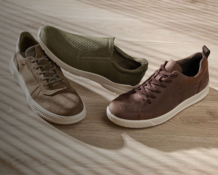Tre par bekväma fritidsskor i olika bruna nyanser - olivgrön, ljusbrun och mörkbrun - med vita sulor på träyta.
